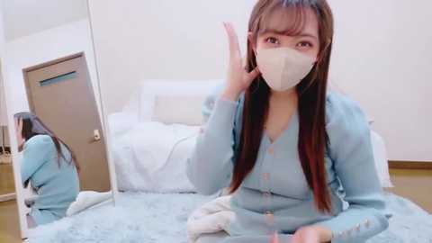 Video of an East Asian woman with long brown hair, wearing a light blue robe, a white mask, and gloves, sitting on a light blue bedspread, gesturing with her right hand, in a minimalist, white room.