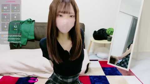 Video of an Asian woman with long brown hair, wearing a black shirt and face mask, sitting on a bed with a colorful quilt. The room has a green jacket hanging on the bed, a plant on a table, and a mirror reflecting another woman.