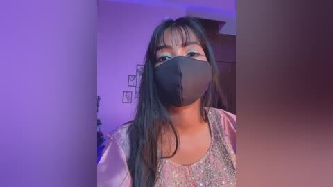 A video of a young Asian woman with long black hair, wearing a black face mask, light blue floral top, and bangs, standing indoors under purple lighting.