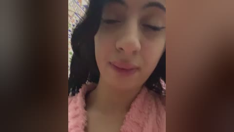 A close-up video of a young Latina woman with medium-brown skin and wavy black hair, wearing a pink fleece robe, smiling softly with closed eyes. Background features colorful, blurred patterned fabric.
