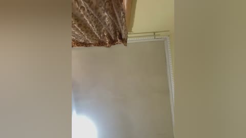Video of a beige wall with a textured, brown, floral-patterned curtain partially hanging over it. The curtain is slightly crooked, revealing a white, framed window with a beige blind partially drawn. The lighting is soft, casting a warm, diffused glow.