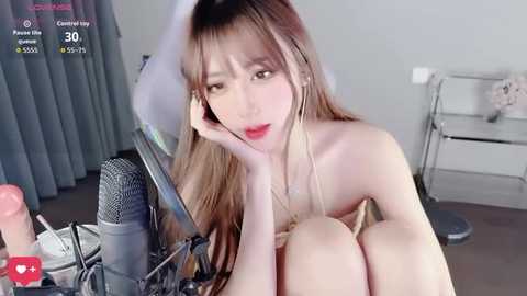 A video of an Asian woman with long, straight brown hair, wearing red lipstick, sitting naked on a chair with a microphone in front of a gray curtain.