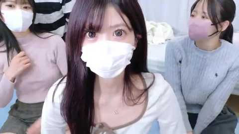 Video of three young Asian women wearing white face masks, sitting on a light blue floor, dressed in casual attire, in a soft-lit room with white walls.