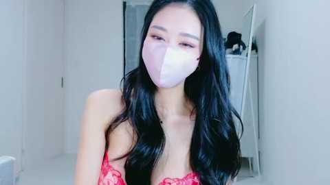 Video of an East Asian woman with long black hair, wearing a pink face mask and red lace lingerie, in a minimalistic, white-walled room.