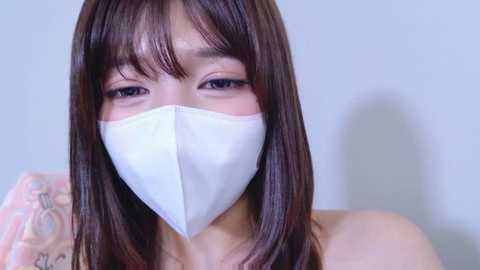 Video of an East Asian woman with long, straight, dark brown hair, wearing a white surgical mask covering her nose and mouth, and a pink floral-patterned top.