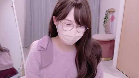Video of a young Asian woman with long brown hair, wearing glasses and a mask, in a bathroom with a pink stool, white tiles, and a blue curtain.