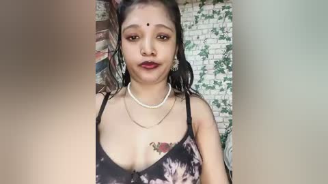 Video of an Indian woman with dark skin, wearing a black floral dress, pearl necklace, and red lipstick, against a brick wall.