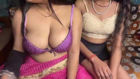 Video of two South Asian women with medium skin tones, one in a purple bra, the other in a beige lace top, sitting on a patterned bed.