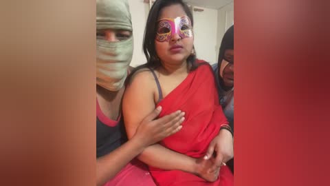 Video of a woman in a red sari with a mask featuring colorful designs, being held by two people, one wearing a surgical mask, in a domestic setting.