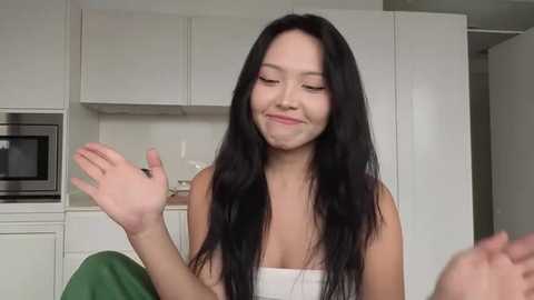 A video of a young Asian woman with long black hair, wearing a white top, smiling and gesturing in a modern kitchen with white cabinets and stainless steel appliances.