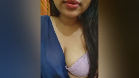 Video of a woman with long black hair, fair skin, and a deep V-neck blue dress, revealing a lavender lace bra with a visible cleavage. The background is blurred, suggesting an indoor setting.