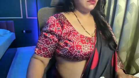 Video of a South Asian woman with long black hair, wearing a red and white patterned blouse and a black and red sari, seated indoors next to a bed with white sheets and dark furniture.
