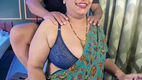 Video of a curvy, dark-skinned woman in a blue floral sari with a lace bra, being massaged by another woman with light brown skin. Background includes a bed, green curtains, and a tiled floor.