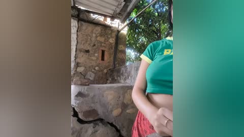 Video of a woman with a medium build, green shirt, and red shorts, standing in front of a rustic stone building with a small window and a cracked floor.