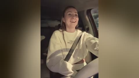 Media: Video of a smiling young woman in a car, wearing a cream sweatshirt and gray jeans, with a shoulder strap across her chest, looking relaxed.