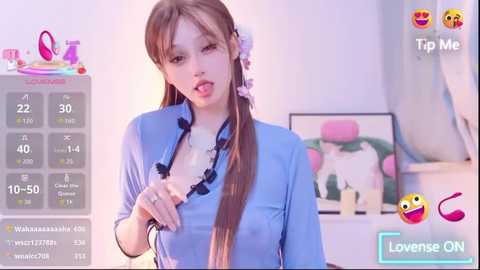 A video of a young woman with long brown hair in a light blue shirt, partially unbuttoned, in a playful, suggestive pose. The background features a pastel-colored room with a framed artwork.