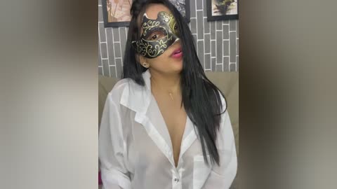 Video of a woman with long black hair, wearing a sheer white blouse, black mask with golden patterns, and pink lipstick, seated in a modern room with gray brick wallpaper and framed artwork.