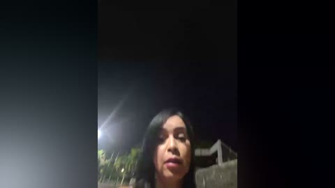 Media: A video of a woman with long, black hair and closed eyes, standing in a dimly lit, dark alley at night, with a bright light source on the left.