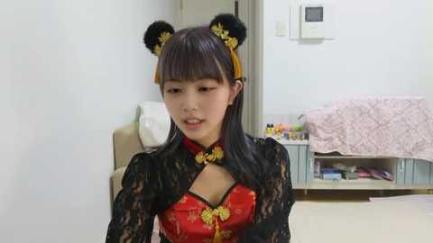 Video of an East Asian woman in a red, black lace, and gold-trimmed Chinese dress with black cat ears, standing indoors. Background shows a bed, desk, and wall with an air conditioner.