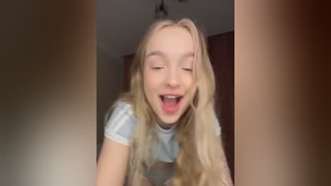 A video captures a blonde, fair-skinned teenage girl with wavy hair, wearing a light blue t-shirt, laughing heartily with her mouth open. The background shows a dimly lit room with wooden furniture and a ceiling light.