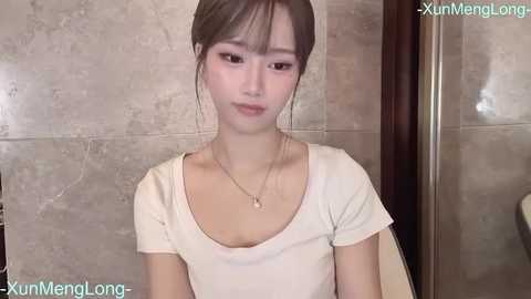 Media: A video of a young East Asian woman with light skin, straight brown hair, and a V-neck white T-shirt, sitting indoors with a beige tiled background and watermarked \"XunMengLong.\
