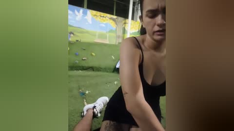 Video of a young woman with fair skin and brown hair, wearing a black tank top, kneeling on a grassy indoor area with colorful climbing toys.