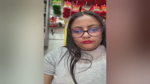 Video of a Latina woman with long dark hair, wearing glasses and a gray T-shirt, standing in a grocery store aisle with red and green snack bags hanging overhead.