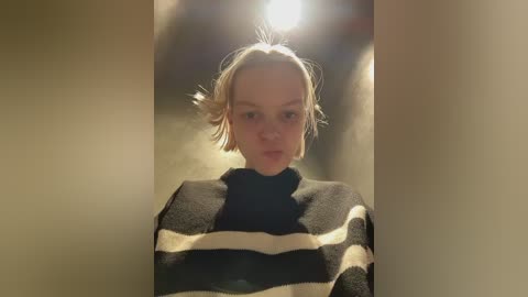 A video of a young, fair-skinned woman with blonde hair, wearing a black and white striped sweater, standing in a dimly lit hallway with a bright light source behind her.