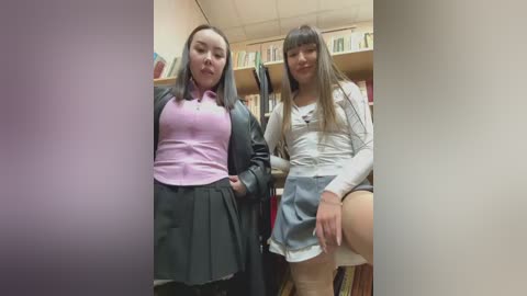 Video of two young Asian women with long hair, one in a pink blouse and black skirt, the other in a white top and grey skirt, standing in a book-filled room.
