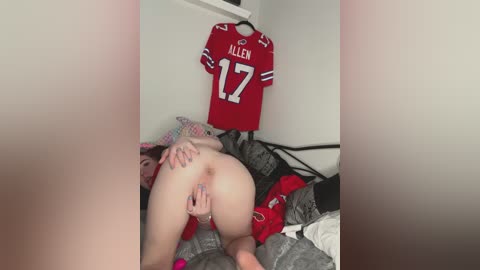 A video of a naked woman with pale skin, dark hair, and a pierced butt, positioned provocatively on a bed, wearing a red \"Allen 17\" jersey, surrounded by scattered clothes and bedding.