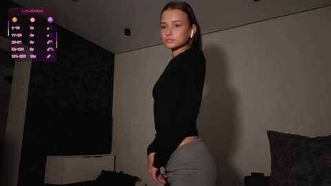 A video of a young woman with light skin, straight brown hair, and a slender physique, wearing a black long-sleeve top and gray pants, standing in a dimly lit bedroom with a flat-screen TV, a dark headboard, and a digital clock on the wall.