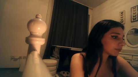 Media: A video shows a young woman with long black hair, wearing a black top, sitting beside a white chess pawn in a dimly lit room with a dark curtain and framed art on the wall.