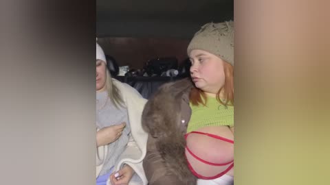 Video of two women in casual clothes; one in a light sweater, the other in a yellow top, both with a dog between them. Background shows a dark room with blurred objects.