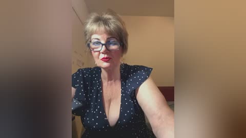 Video of a middle-aged woman with short, light brown hair, wearing glasses and a black polka-dotted blouse with a plunging neckline, standing indoors with beige walls and a red object partially visible in the background.