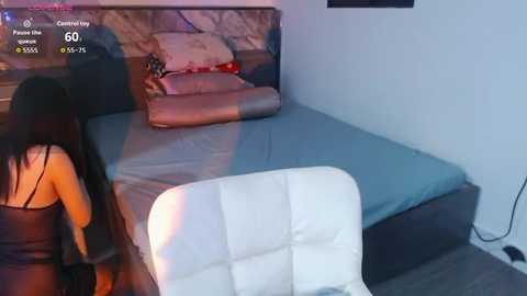 A video of a modern bedroom with a woman in a black dress and a man in a white chair. The bed features a gray duvet and pillows.