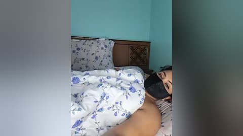 A video captures a woman, likely of Asian descent, lying on a bed with white sheets featuring blue floral patterns. She has dark hair, wears a black face mask, and appears partially covered by the sheets, with a wooden headboard and turquoise wall visible in the background.