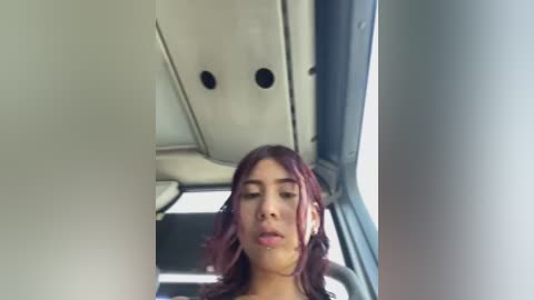 Video of a young woman with wavy, dark red hair, wearing a black top, seated in a modern airplane cabin with white seats and windows.