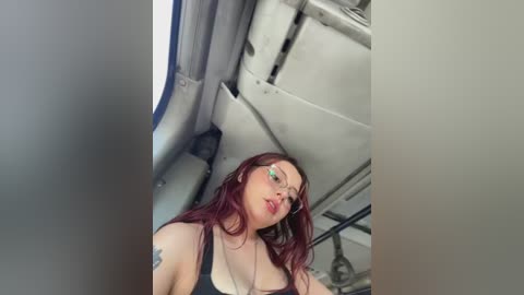 Video of a plus-size woman with long red hair, wearing glasses, a black tank top, and a tattoo, taken in an airplane bathroom, with a blurry toilet in the background.