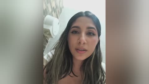 Video of a Latina woman with long, wavy, dark hair, wearing a white shirt, standing in a bathroom with beige tiles, holding a white paper towel roll.