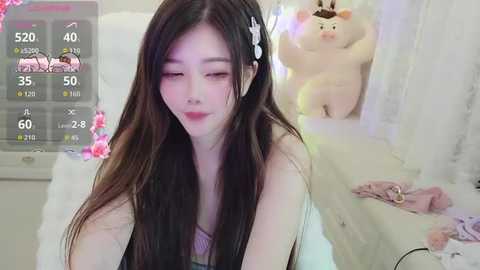 Video of a young East Asian woman with long, straight black hair, wearing a white top, smiling in a cozy, pink-themed room with plush toys and a screen displaying game stats.