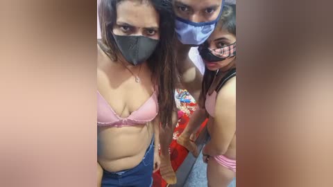 Video of three women, wearing face masks, in pink and blue lingerie, holding hands in a close, intimate pose.