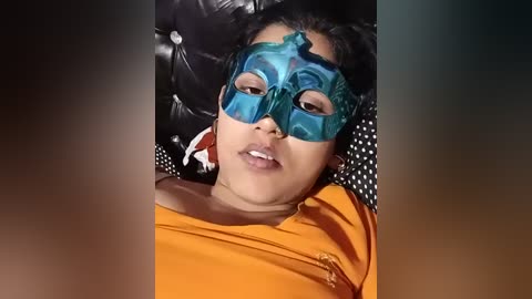 Video of a young child with medium brown skin, wearing a teal Venetian mask and a bright orange shirt, lying on a tufted black leather couch with a polka-dotted pillow.