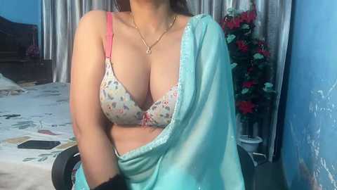 Media: Video of a woman in a floral bra, teal shawl, and gold necklace, seated on a chair in a room with a blue wall, floral arrangement, and white carpet.