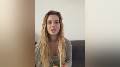 Video of a young Caucasian woman with long, wavy blonde hair, wearing a grey top, sitting on a grey couch against a plain white wall.