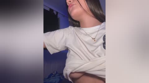 A video of a woman lifting her white T-shirt to expose her left breast, revealing small, light brown nipples. She has long, straight dark hair and wears a thin necklace. Background includes a dark curtain and a bed with blue sheets.