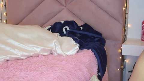 Video of a cozy bedroom corner with a plush, pink quilted headboard, a white pillow, and a navy blue blanket partially draped over a pink, fluffy blanket on a bed.