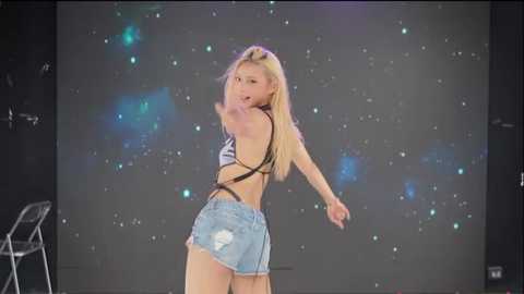 Video of a blonde, slim, light-skinned woman in a black bra and distressed denim shorts dancing on stage, background features a starry sky projection.