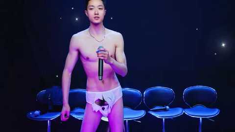 Video of a shirtless Asian man with fair skin, wearing white briefs and a necklace, singing into a microphone against a dark background with blue lighting and modern chairs.