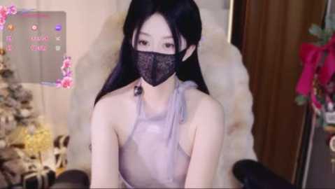Video of an East Asian woman with long black hair, wearing a gray, sheer, halterneck top, black mask, and floral wallpaper.