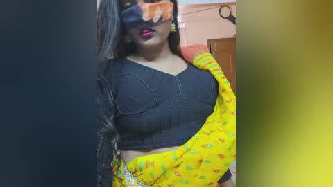 Video of a woman with medium skin tone, wearing a black crop top and yellow sari with colorful patterns, covering her face with an orange mask.